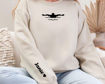 Peut inclure: Sweat-shirt beige avec un motif de nageur noir sur la poitrine. La manche gauche porte le nom "Janie" en noir avec un cœur. Le sweat-shirt à col rond est de couleur neutre.