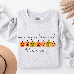 Puede incluir: Una sudadera gris claro con las palabras "occupational therapy" en una fuente cursiva. Debajo del texto hay una fila de siete calabazas de dibujos animados en naranja y amarillo. Un sombrero marrón y hojas de palma secas en el fondo.