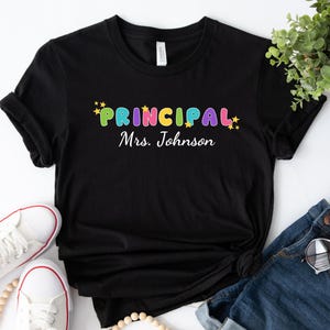 Op de afbeelding: Zwart T-shirt met de tekst "PRINCIPAL Mevrouw Johnson" in kleurrijke letters met sterren aan beide kanten.