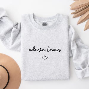 Op de afbeelding: Een lichtgrijs sweatshirt met de tekst "admin team" in zwart script en een smiley. De mouwen zijn opgerold. Een beige hoed en een gedroogde plant staan op de achtergrond. Het sweatshirt ligt plat.