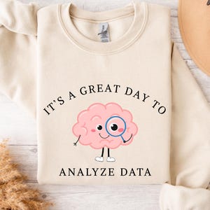 Puede incluir: Sudadera de color crema con el texto "IT'S A GREAT DAY TO ANALYZE DATA". El gráfico presenta un cerebro rosa con una lupa. La sudadera tiene cuello redondo y mangas largas.