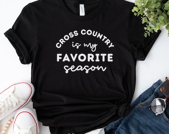 Może przedstawiać: Czarny t-shirt z białym napisem "Cross Country is my FAVORITE season". Koszulka jest połączona z dżinsowymi szortami, białymi trampkami i okularami przeciwsłonecznymi. Szereg drewnianych koralików i zieleń uzupełniają swobodny zestaw.