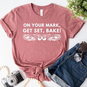 May include: A dusty rose t-shirt with rolled sleeves and a knotted hem. The shirt features the text "ON YOUR MARK, GET SET, BAKE!" in white script. A pair of denim shorts, sunglasses, and a camera are also visible.