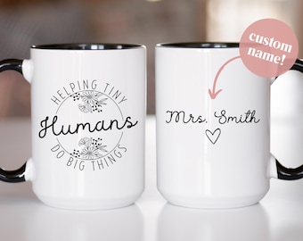May include: Two white ceramic mugs with black handles and rims. One mug has the text "Helping Tiny Humans Do Big Things" in a floral wreath. The other mug has the text "Mrs. Smith" with a heart symbol. The mugs are for custom names.