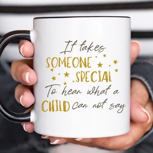 May include: White ceramic mug with a black handle and rim, featuring the text "It takes SOMEONE SPECIAL To hear what a CHILD can not say" in gold and black lettering. The mug is held by a person.