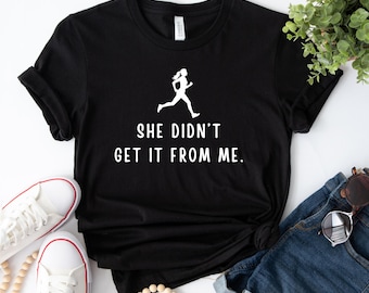 Może przedstawiać: Czarny t-shirt z białym napisem i grafiką biegacza. Tekst brzmi "SHE DIDN'T GET IT FROM ME." Obraz zawiera białe trampki, dżinsowe szorty i okulary przeciwsłoneczne, sugerując sportowy, swobodny motyw.