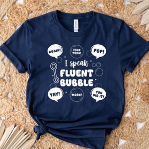 May include: Navy blue t-shirt with the text "I speak FLUENT BUBBLE" in white bubble letters. The shirt also features speech bubbles with words like "AGAIN!", "POP!", and "YAY!". The shirt is displayed on a woven, beige surface.