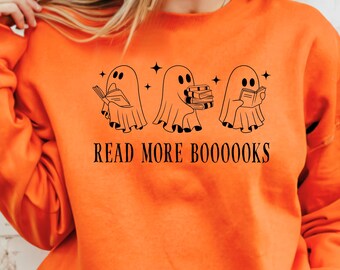 May include: Orange sweatshirt with a black graphic of three ghost figures reading books. The text "READ MORE BOOOOOKS" is printed below the ghosts. The sweatshirt has long sleeves and a crew neck.