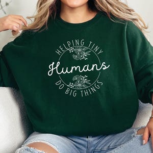 May include: A dark green sweatshirt featuring the text "Helping Tiny Humans Do Big Things" in a white, circular design with floral accents. The sweatshirt is a solid colour and has long sleeves.
