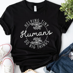 Puede incluir: Camiseta negra con el texto "Helping Tiny Humans Do Big Things" en un diseño floral blanco. La imagen incluye zapatillas blancas, pantalones cortos vaqueros y un collar de cuentas.