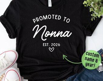 May include: Black t-shirt with the text "PROMOTED TO Nonna EST. 2026" in white script and a small heart. The shirt is styled with white sneakers, denim shorts, and a beaded necklace.