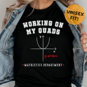 May include: Black t-shirt with the text "WORKING ON MY QUADS" in white, along with a mathematical graph and the equation "y=ax²+bx+c". Below the equation is the text "MATHLETICS DEPARTMENT". A yellow circle with "UNISEX FIT!" is in the upper right.