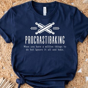 May include: Navy blue t-shirt with the text "PROCRASTIBAKING" in white, along with crossed rolling pins and wheat sprigs. The shirt also includes the phrase "When you have a million things to do but ignore it all and bake."