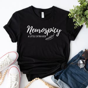 May include: Black t-shirt with the text "Neurospicy" in a cursive font, with "A LITTLE EXTRA KICK" below and a chili pepper graphic. The shirt is styled with white sneakers, denim shorts, and sunglasses.