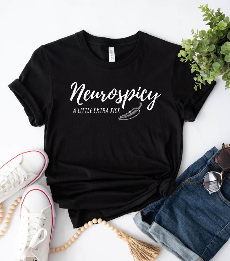 May include: Black t-shirt with the text "Neurospicy" in a cursive font, with "A LITTLE EXTRA KICK" below and a chili pepper graphic. The shirt is styled with white sneakers, denim shorts, and sunglasses.