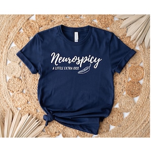 May include: Navy blue t-shirt with the text "Neurospicy" in a cursive font, accompanied by "A LITTLE EXTRA KICK" and a chili pepper graphic. The shirt is knotted at the bottom.