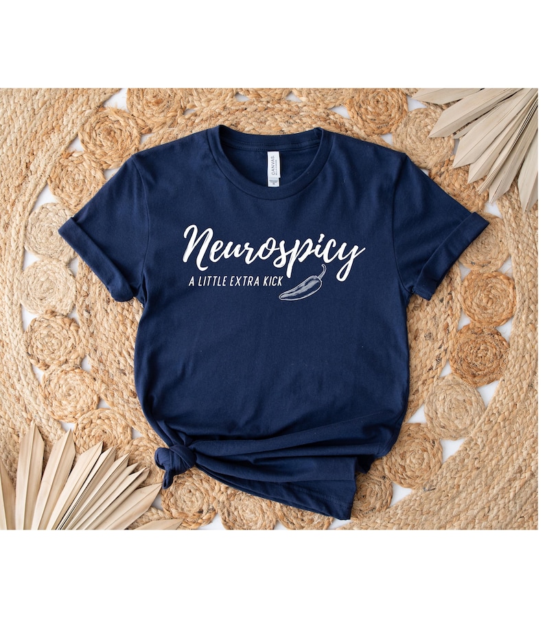 May include: Navy blue t-shirt with the text "Neurospicy" in a cursive font, accompanied by "A LITTLE EXTRA KICK" and a chili pepper graphic. The shirt is knotted at the bottom.