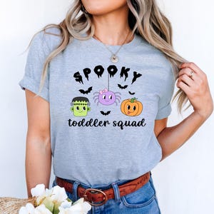 Peut inclure: T-shirt gris clair avec un motif sur le th&egrave;me d'Halloween. Le motif comprend le mot "SPOOKY" au-dessus d'une araign&eacute;e, d'une citrouille et du monstre de Frankenstein. Le texte "toddler squad" est en dessous. Le t-shirt a des manches courtes.