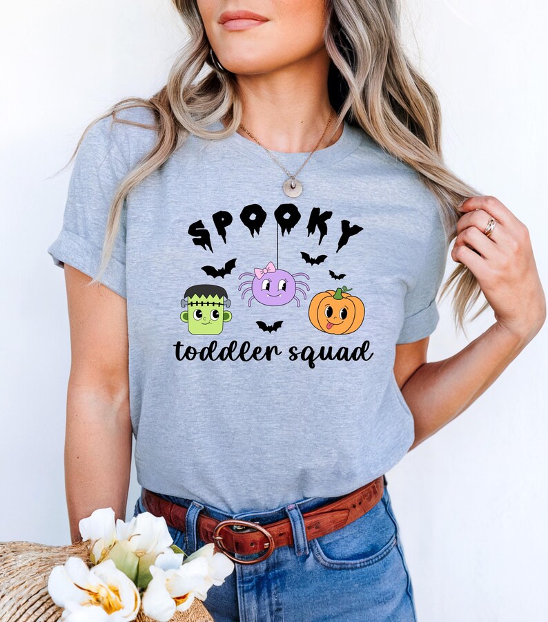 Peut inclure: T-shirt gris clair avec un motif sur le th&egrave;me d'Halloween. Le motif comprend le mot "SPOOKY" au-dessus d'une araign&eacute;e, d'une citrouille et du monstre de Frankenstein. Le texte "toddler squad" est en dessous. Le t-shirt a des manches courtes.