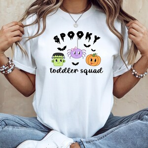 Peut inclure: T-shirt blanc avec un motif sur le th&egrave;me d'Halloween. Le motif comprend le texte "Spooky toddler squad" avec des illustrations d'un monstre de Frankenstein, d'une araign&eacute;e, d'une citrouille et de chauves-souris.
