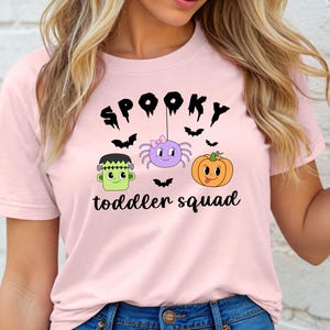Peut inclure: T-shirt rose clair avec un motif sur le th&egrave;me d'Halloween. Le mot "Spooky" est au-dessus d'une araign&eacute;e violette, d'une citrouille orange et d'un monstre de Frankenstein vert. Le texte "toddler squad" est en dessous.