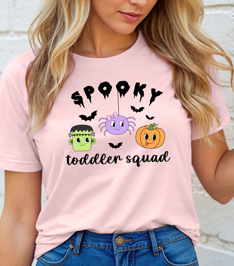 Peut inclure: T-shirt rose clair avec un motif sur le th&egrave;me d'Halloween. Le mot "Spooky" est au-dessus d'une araign&eacute;e violette, d'une citrouille orange et d'un monstre de Frankenstein vert. Le texte "toddler squad" est en dessous.