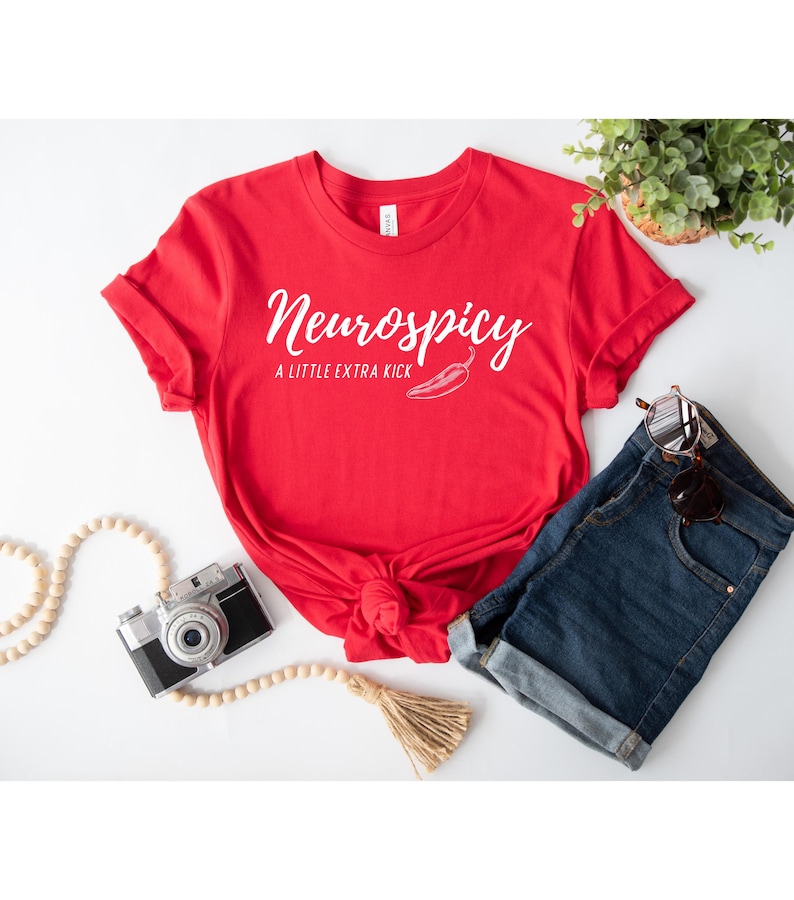 May include: A red t-shirt with the text "Neurospicy" in a cursive font, along with "A Little Extra Kick" and a chili pepper graphic. The shirt is styled with denim shorts, sunglasses, and a vintage camera.