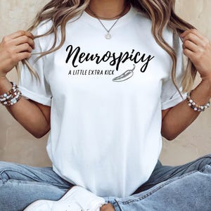 May include: White t-shirt with the text "Neurospicy: A Little Extra Kick" in black script, accompanied by a chili pepper illustration. The shirt is a classic crew neck style.