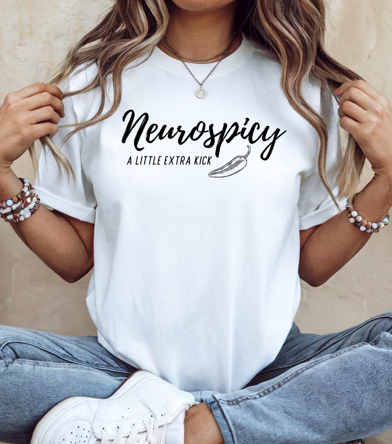 May include: White t-shirt with the text "Neurospicy: A Little Extra Kick" in black script, accompanied by a chili pepper illustration. The shirt is a classic crew neck style.