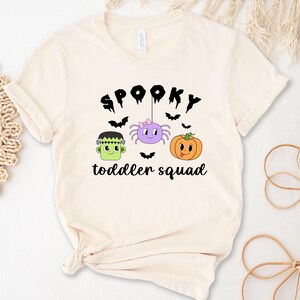 Peut inclure: T-shirt cr&egrave;me avec l'inscription noire "Spooky toddler squad". Le motif comprend un Frankenstein vert, une araign&eacute;e violette avec un n&oelig;ud rose et une citrouille orange. Des chauves-souris noires sont dispers&eacute;es.