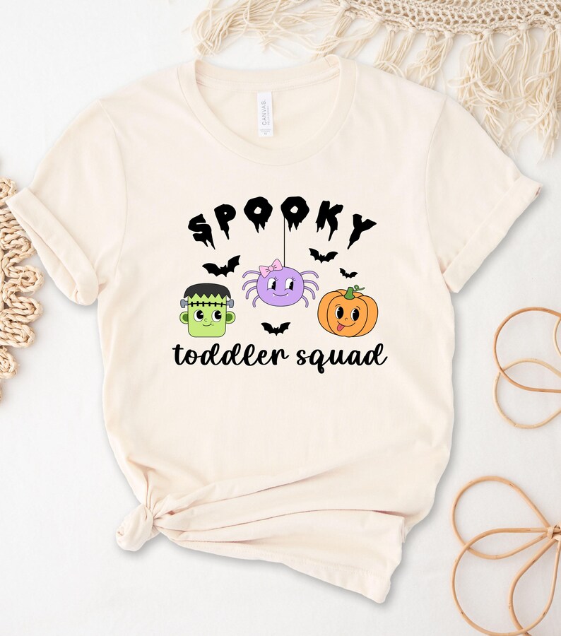 Peut inclure: T-shirt cr&egrave;me avec l'inscription noire "Spooky toddler squad". Le motif comprend un Frankenstein vert, une araign&eacute;e violette avec un n&oelig;ud rose et une citrouille orange. Des chauves-souris noires sont dispers&eacute;es.
