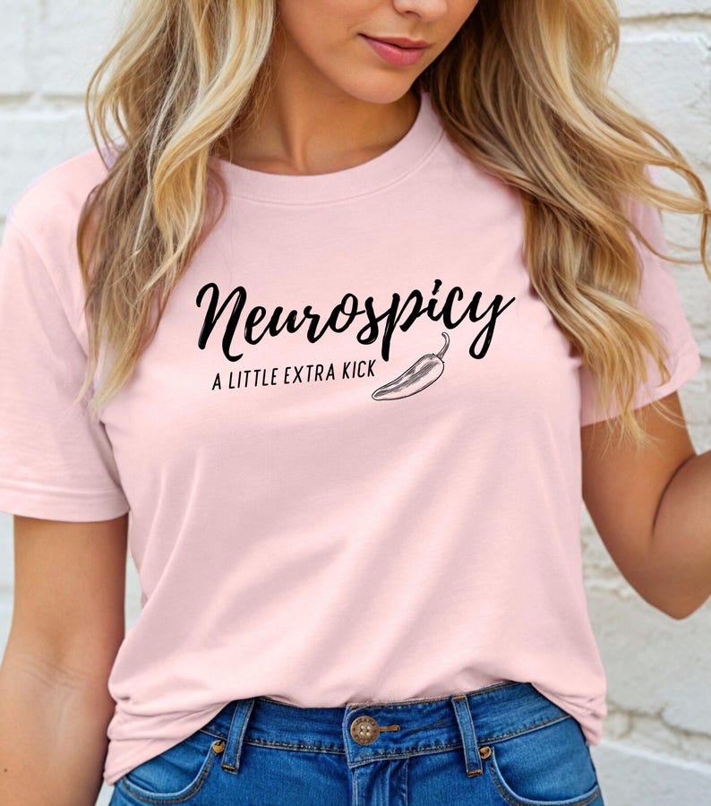 May include: A light pink t-shirt with the text "Neurospicy" in a black script font, accompanied by the phrase "A LITTLE EXTRA KICK" and a chili pepper illustration. The shirt is paired with blue jeans.