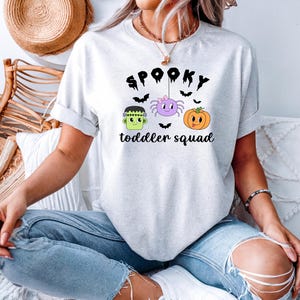Peut inclure: T-shirt gris clair avec un motif sur le th&egrave;me d'Halloween. Le motif comprend le mot "Spooky" au-dessus d'un dessin anim&eacute; de Frankenstein, d'une araign&eacute;e et d'une citrouille. Le texte "toddler squad" est en dessous. Le t-shirt est en mati&egrave;re douce.