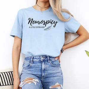 May include: Light blue t-shirt with the text "Neurospicy" in a script font, accompanied by "A LITTLE EXTRA KICK" and a chili pepper graphic. The shirt is paired with blue denim jeans.