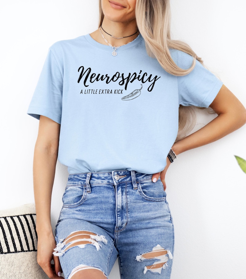 May include: Light blue t-shirt with the text "Neurospicy" in a script font, accompanied by "A LITTLE EXTRA KICK" and a chili pepper graphic. The shirt is paired with blue denim jeans.