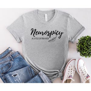 May include: A light gray t-shirt with the text "Neurospicy" in a cursive font, accompanied by "A LITTLE EXTRA KICK" and a chili pepper graphic. The shirt is paired with blue jeans and white sneakers.