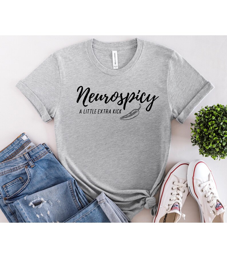 May include: A light gray t-shirt with the text "Neurospicy" in a cursive font, accompanied by "A LITTLE EXTRA KICK" and a chili pepper graphic. The shirt is paired with blue jeans and white sneakers.