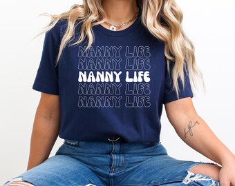 Może przedstawiać: Granatowa koszulka z krótkim rękawem z napisem NANNY LIFE wydrukowanym pięć razy w stosie, falowaną białą czcionką. Środkowa linia tekstu jest w kolorze białym, podczas gdy pozostałe cztery linie mają biały kontur, tworząc powtarzający się wzór.