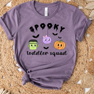 Peut inclure: Un t-shirt violet avec l'inscription "SPOOKY toddler squad" en noir. Le motif comprend un Frankenstein vert, une araign&eacute;e violette avec un n&oelig;ud rose et une citrouille orange. Des chauves-souris noires sont dispers&eacute;es.