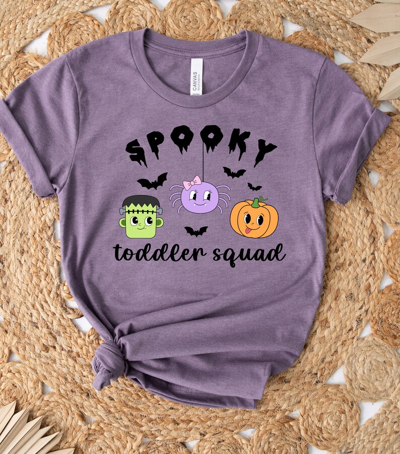 Peut inclure: Un t-shirt violet avec l'inscription "SPOOKY toddler squad" en noir. Le motif comprend un Frankenstein vert, une araign&eacute;e violette avec un n&oelig;ud rose et une citrouille orange. Des chauves-souris noires sont dispers&eacute;es.