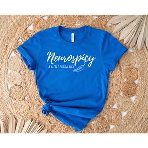 May include: A royal blue t-shirt with the text "Neurospicy" in a white script font, accompanied by the phrase "A Little Extra Kick" and a chili pepper illustration. The shirt is knotted at the bottom and rests on a woven, beige surface.