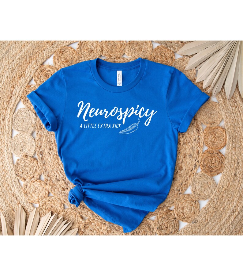 May include: A royal blue t-shirt with the text "Neurospicy" in a white script font, accompanied by the phrase "A Little Extra Kick" and a chili pepper illustration. The shirt is knotted at the bottom and rests on a woven, beige surface.