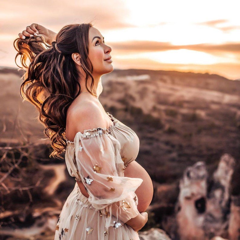 May include: A woman in a beige off-the-shoulder maternity dress with sheer sleeves adorned with star-shaped embellishments. She is outdoors, looking up, with her hand in her hair, against a backdrop of a sunset and rolling hills.