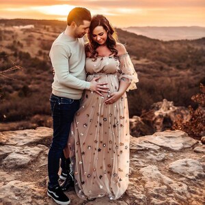 May include: A couple embraces outdoors at sunset. The woman wears a beige off-the-shoulder maternity dress adorned with star-shaped sequins. The man wears a light-colored sweater and dark jeans. They stand on a rocky outcrop with a scenic mountain backdrop.