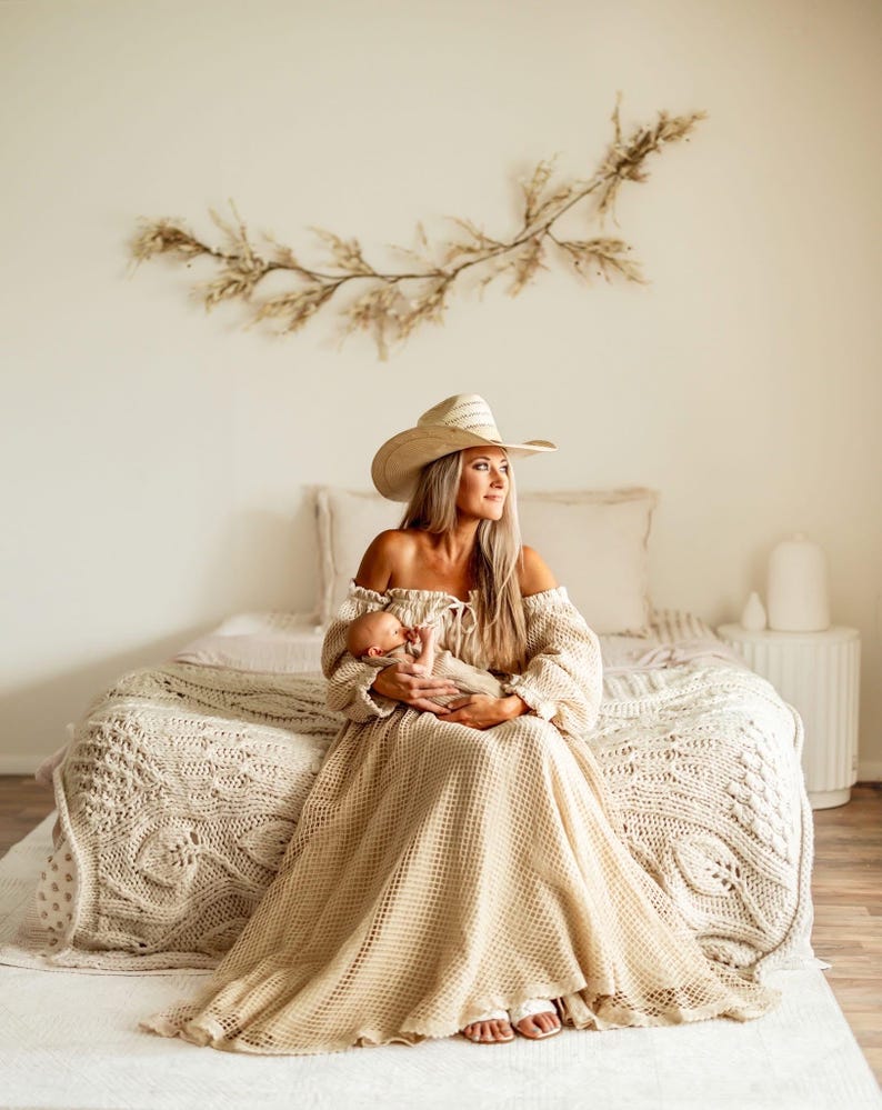 May include: A woman wearing a beige off-the-shoulder dress and a cowboy hat, holding a baby. The dress has long sleeves and a flowing skirt. The bed has a cream-colored knitted blanket. A decorative floral arrangement hangs on the wall.