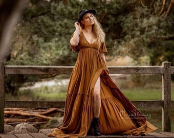 May include: A woman wearing a long, flowing, rust-colored dress with a high slit, standing on a wooden bridge. She wears a black hat and boots. The background features a blurred forest scene with natural light.