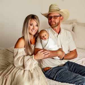 May include: A family portrait featuring a mother in a beige off-the-shoulder dress, a father in a cowboy hat, and a newborn baby wrapped in a white blanket. The family is seated on a bed with a cream-colored knitted blanket.