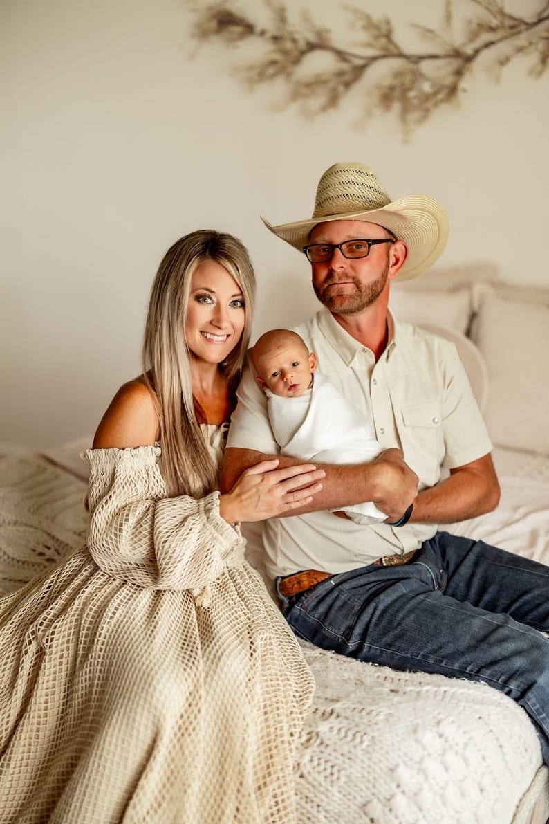 May include: A family portrait featuring a mother in a beige off-the-shoulder dress, a father in a cowboy hat, and a newborn baby wrapped in a white blanket. The family is seated on a bed with a cream-colored knitted blanket.