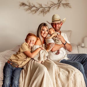 May include: A family portrait featuring a mother, father, and two children. The father wears a cowboy hat and glasses, holding a newborn baby. The older child is smiling and hugging a blanket. The family is sitting on a bed.