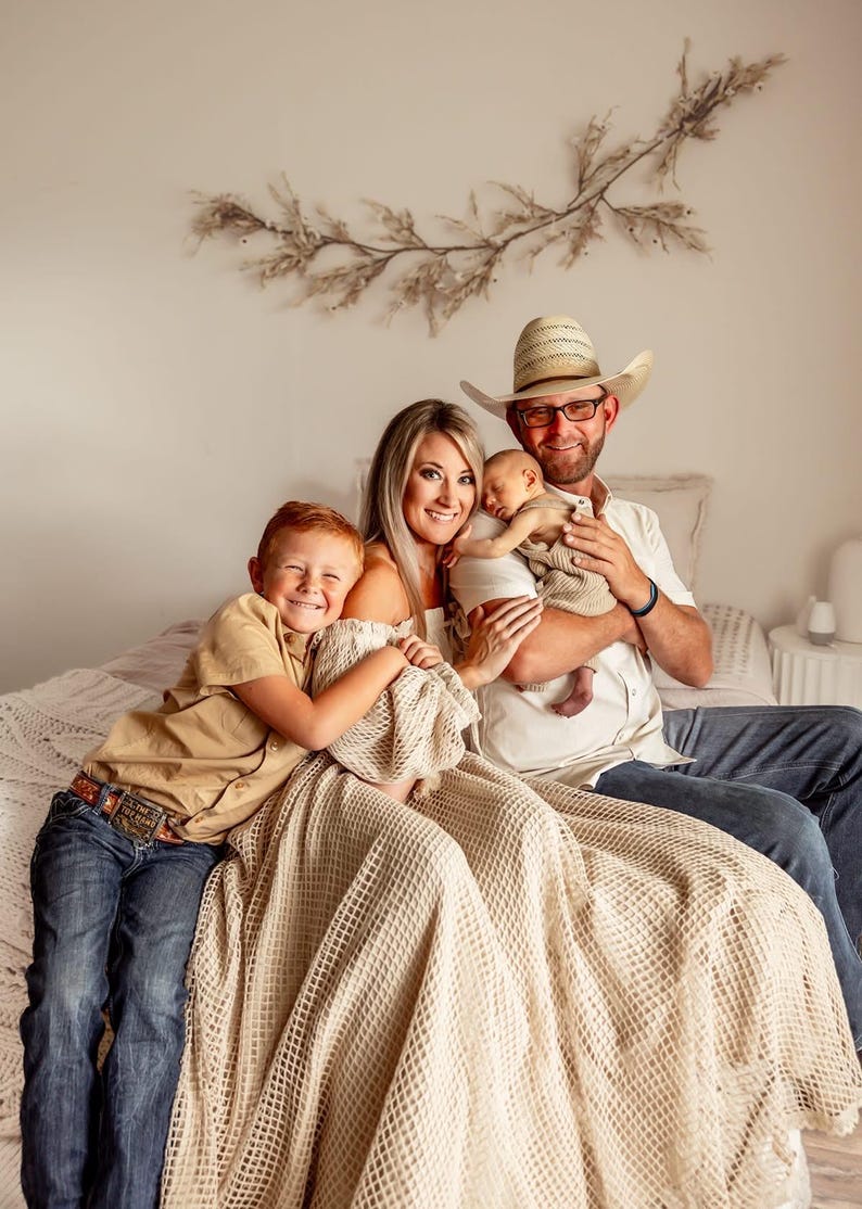 May include: A family portrait featuring a mother, father, and two children. The father wears a cowboy hat and glasses, holding a newborn baby. The older child is smiling and hugging a blanket. The family is sitting on a bed.